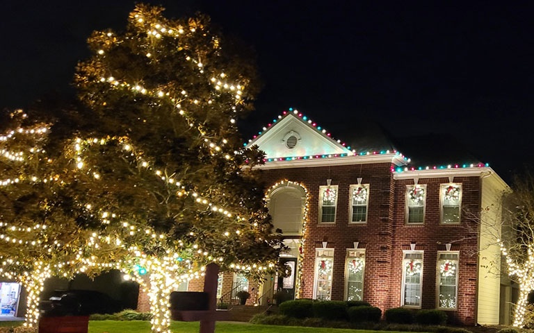 Brick home with colorful roofline lights and trees wrapped in warm white lights